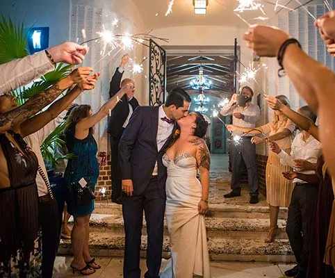 A newlywed couple kisses on stone steps as guests holding sparklers celebrate around them at night. The bride wears a strapless dress and the groom is in a suit. The scene is festive and joyful.
