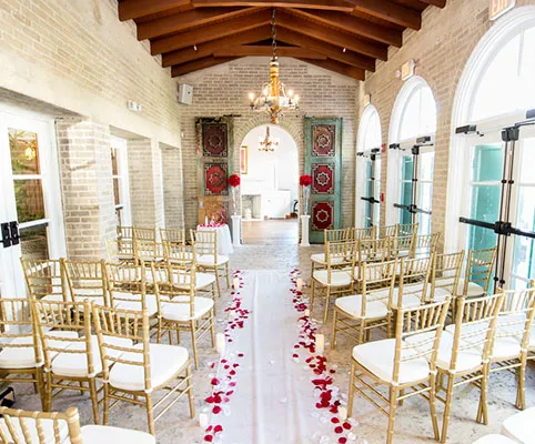 A sunlit indoor wedding ceremony setup with gold chairs arranged in rows, a white aisle runner with red rose petals, and chandeliers hanging from a wooden ceiling. Large arched windows line the right wall.
