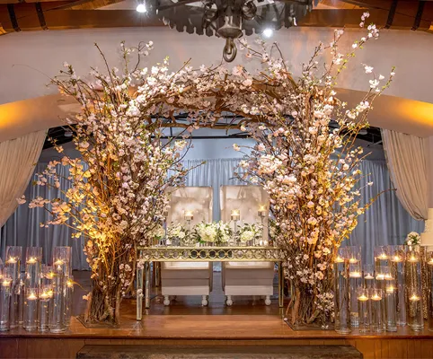 A beautifully decorated wedding stage features two white chairs beneath an archway of blooming branches and flowers, surrounded by tall glass vases filled with lit candles. Soft drapes frame the elegant, romantic setting.