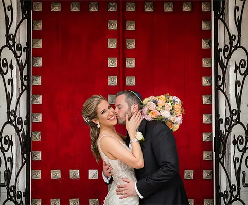 A groom kisses his smiling bride on the cheek as they stand in front of ornate red doors. The bride holds a colorful bouquet and wears a white dress, while the groom is in a black suit.