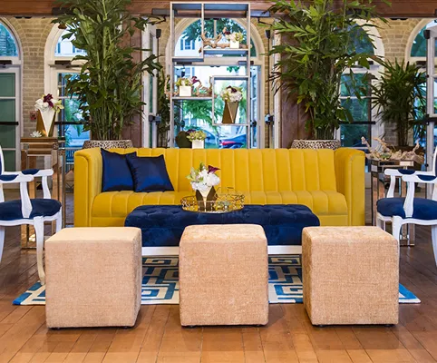 A modern living room with a yellow sofa, blue and gold accents, three beige ottomans, lush green plants, and large windows letting in natural light.