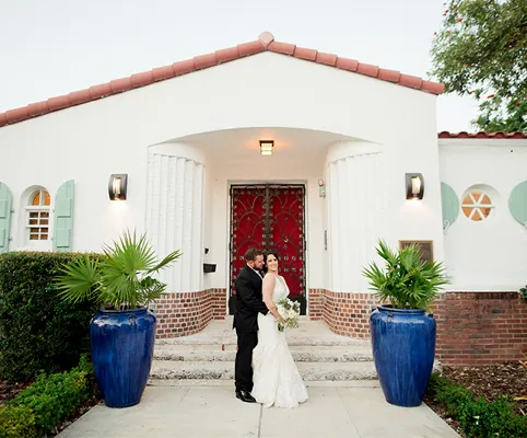A bride and groom pose together in front of a white building with a red door, flanked by large blue planters holding green plants. The bride holds a bouquet and the groom hugs her.