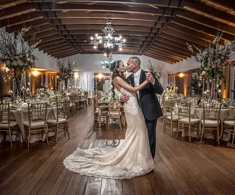 A bride and groom share a romantic kiss while dancing in the center of an elegant, candlelit wedding reception hall decorated with flowers and gold chairs.