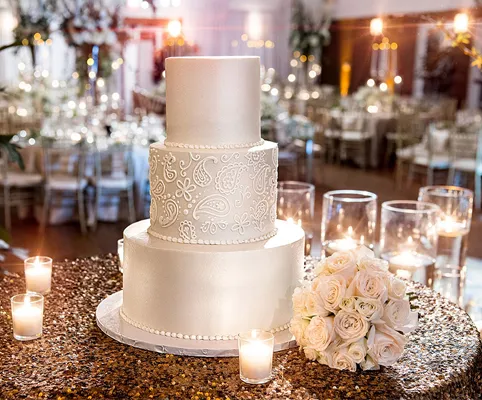 A three-tiered white wedding cake with floral designs is displayed on a sparkling table, surrounded by lit candles and a bouquet of white roses in an elegant reception hall.