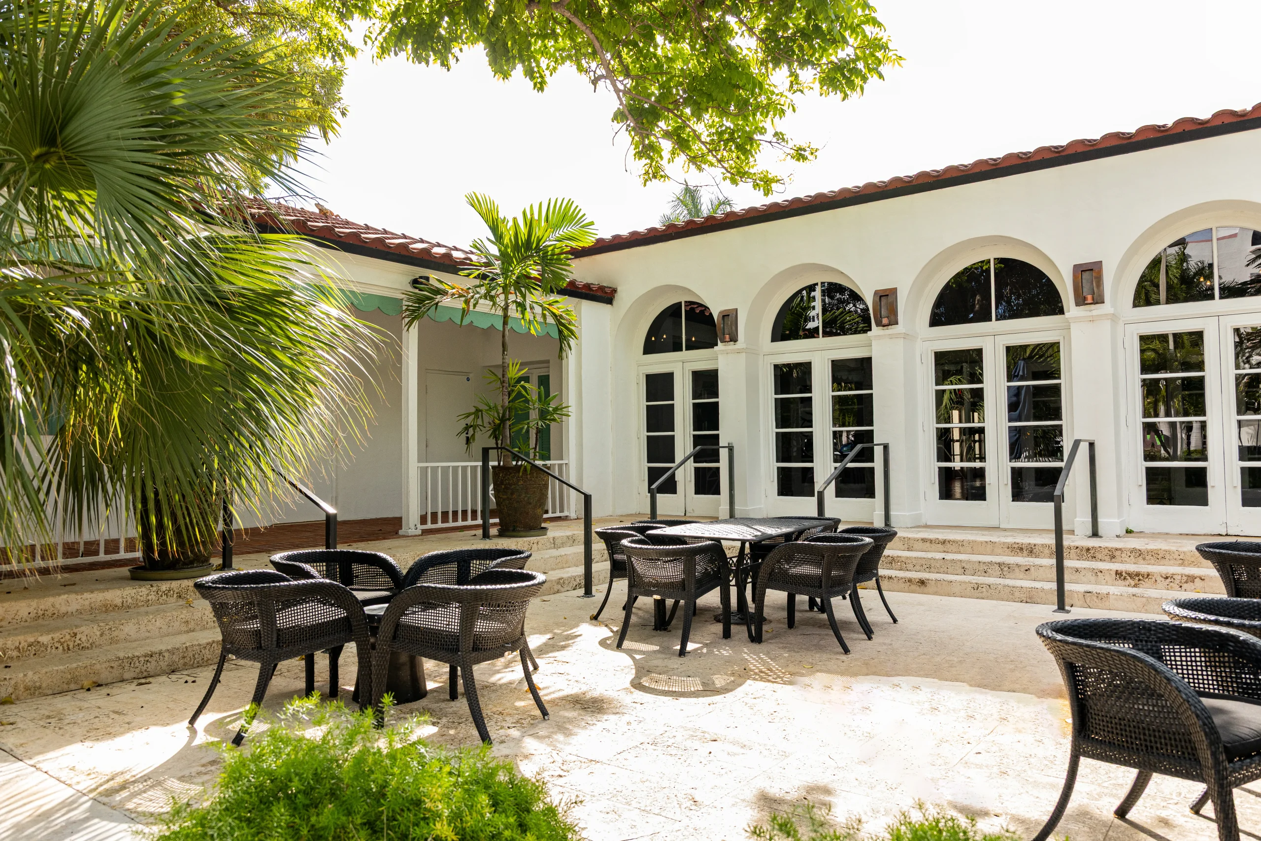 Outdoor patio with several black wicker tables and chairs, surrounded by tropical plants and trees. The building in the background has large arched windows and white walls with a red tiled roof.