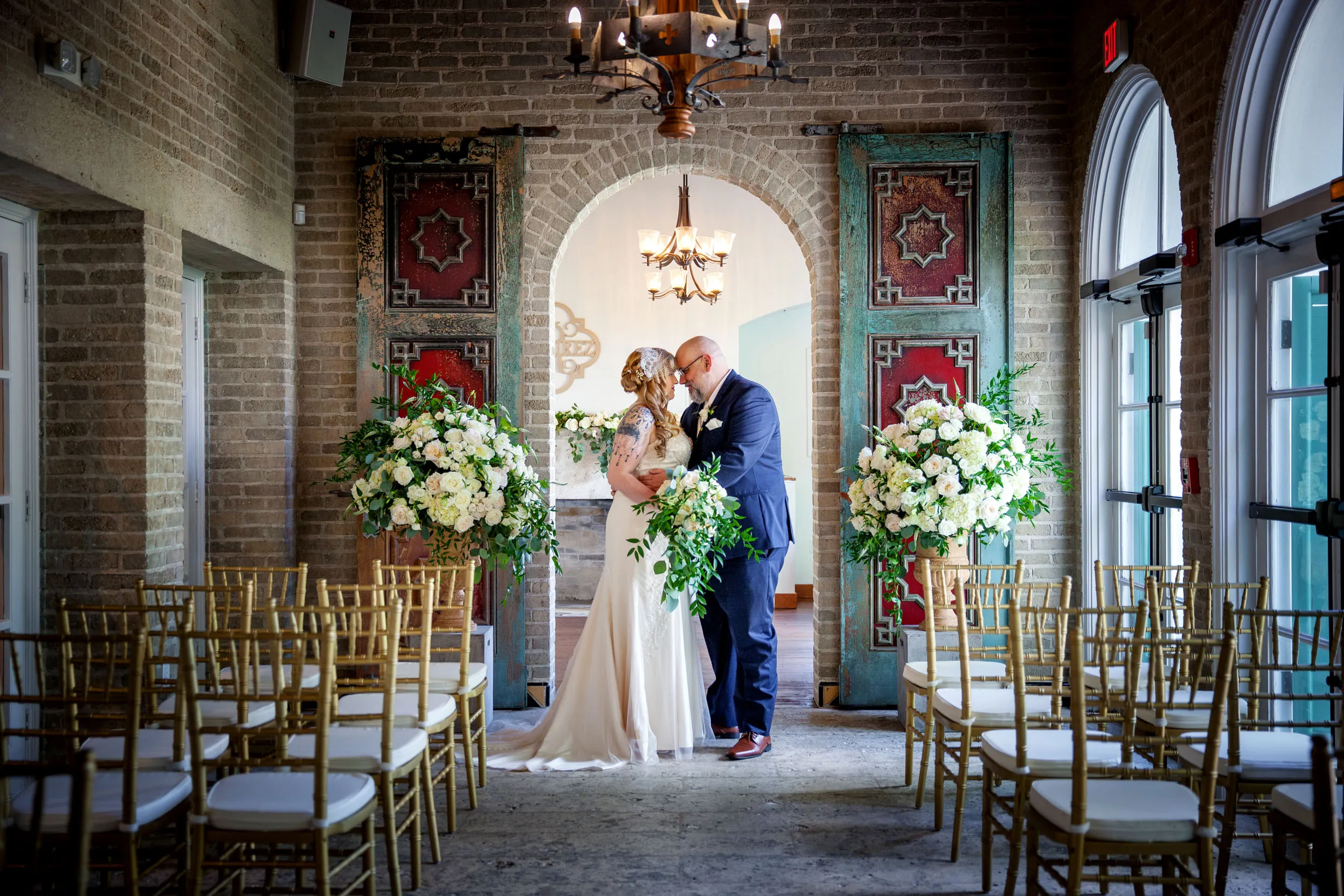 A bride and groom stand closely together in an elegant indoor wedding venue, surrounded by large bouquets of white flowers and gold chairs, beneath a decorative archway with rustic doors and a chandelier overhead.