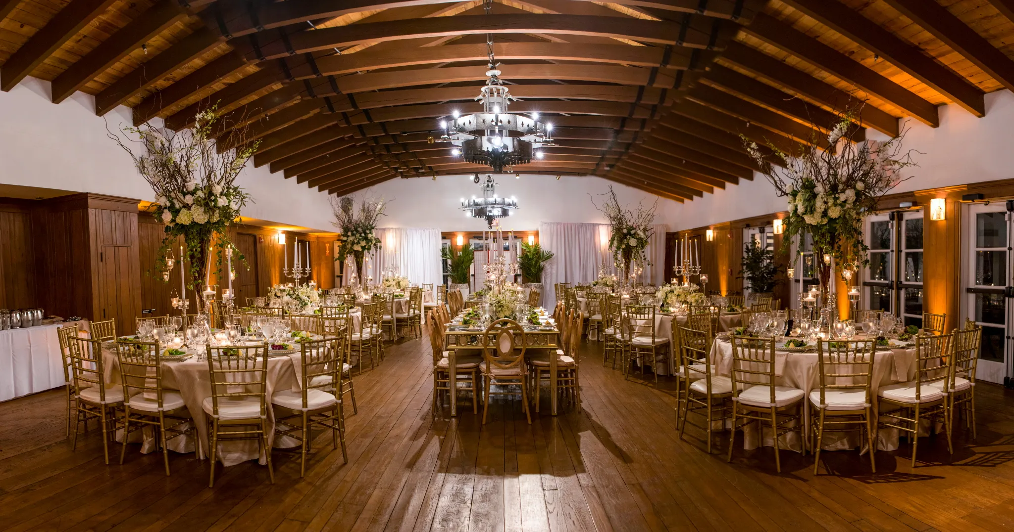 Elegant banquet hall with wooden floors and ceiling beams, decorated for an event. Round tables with gold chairs, floral centerpieces, candles, and soft lighting create a warm, festive atmosphere.