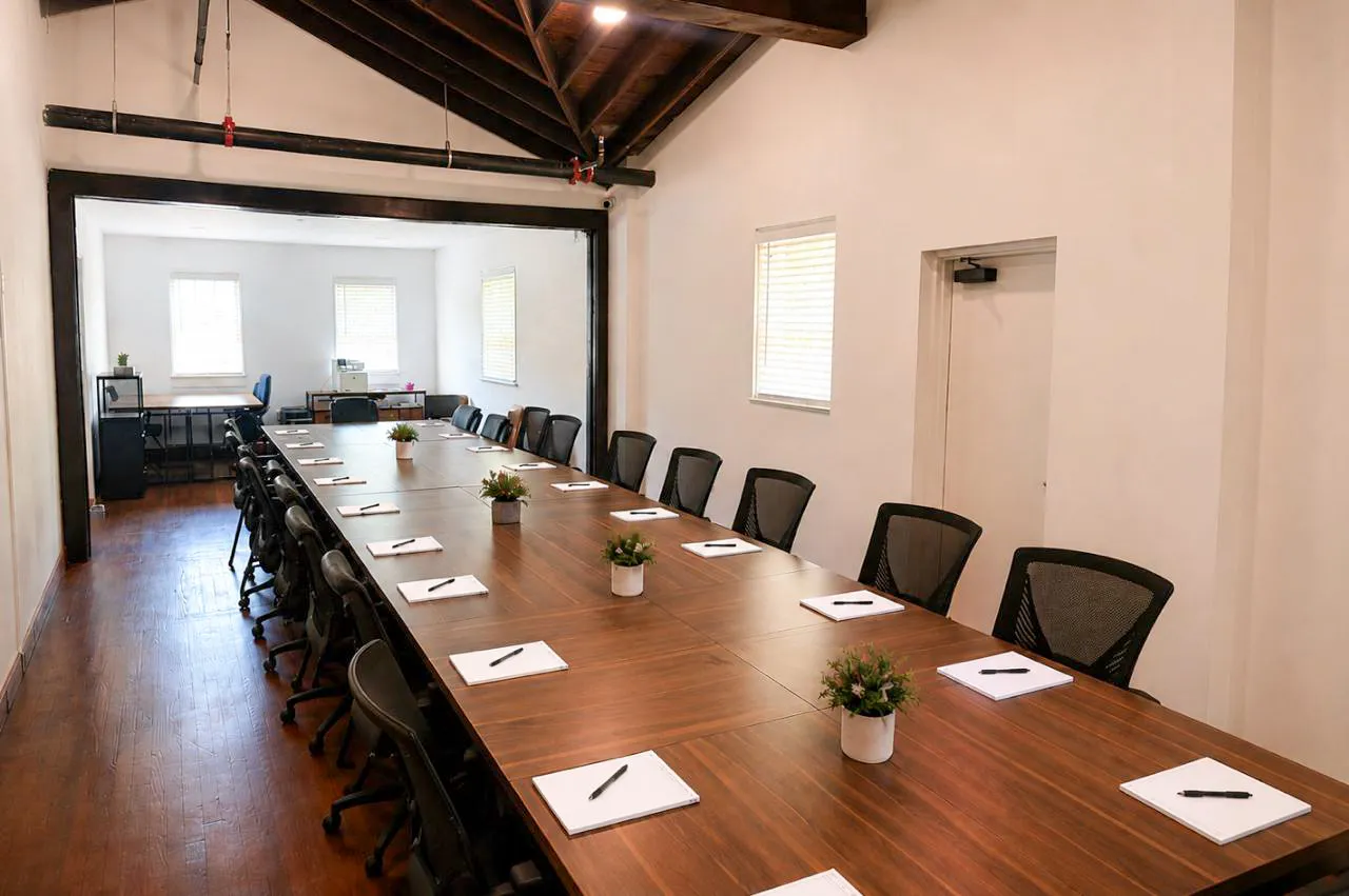 A long conference table with black chairs, notepads, pens, and small potted plants arranged neatly. The room has wooden floors, white walls, windows, and exposed wooden beams on the ceiling.