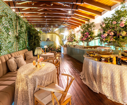 Elegant indoor dining area with wooden floors, beamed ceiling, round tables with floral arrangements, cushioned seating, and a buffet table adorned with decorative cloth and large vases of flowers. Greenery decorates the walls.