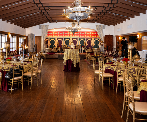 A large banquet hall with wooden floors, chandeliers, round tables covered in ornate gold and burgundy tablecloths, and gold chairs. The stage has decorative drapes and pillows, with lavish decor throughout the room.