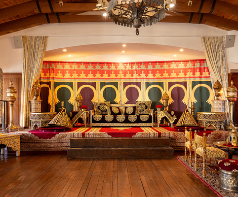 A lavish, Moroccan-inspired room featuring ornate gold-trimmed furniture, vibrant patterned textiles, and a colorful backdrop with arch designs, all set on a raised wooden platform with decorative rugs.