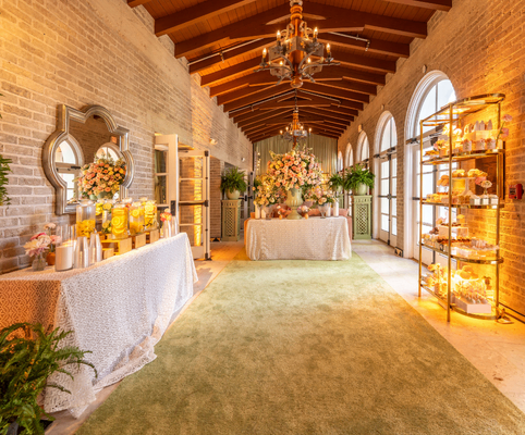 A bright, elegant hallway with arched windows, a green carpet, and wooden ceiling beams. Tables display flowers, food, and drinks, while shelves hold more treats. Warm lighting adds a cozy atmosphere.